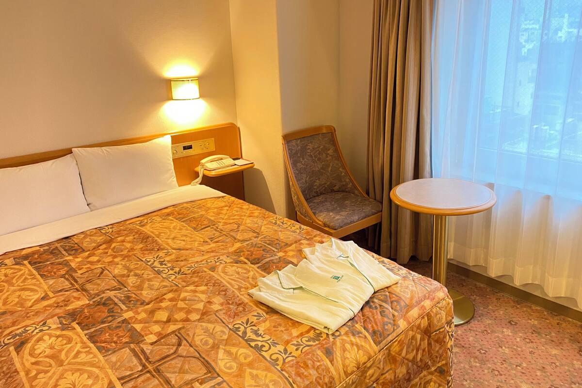 A hotel room with a bed, a chair, a small table, a bedside lamp, and a window with curtains.