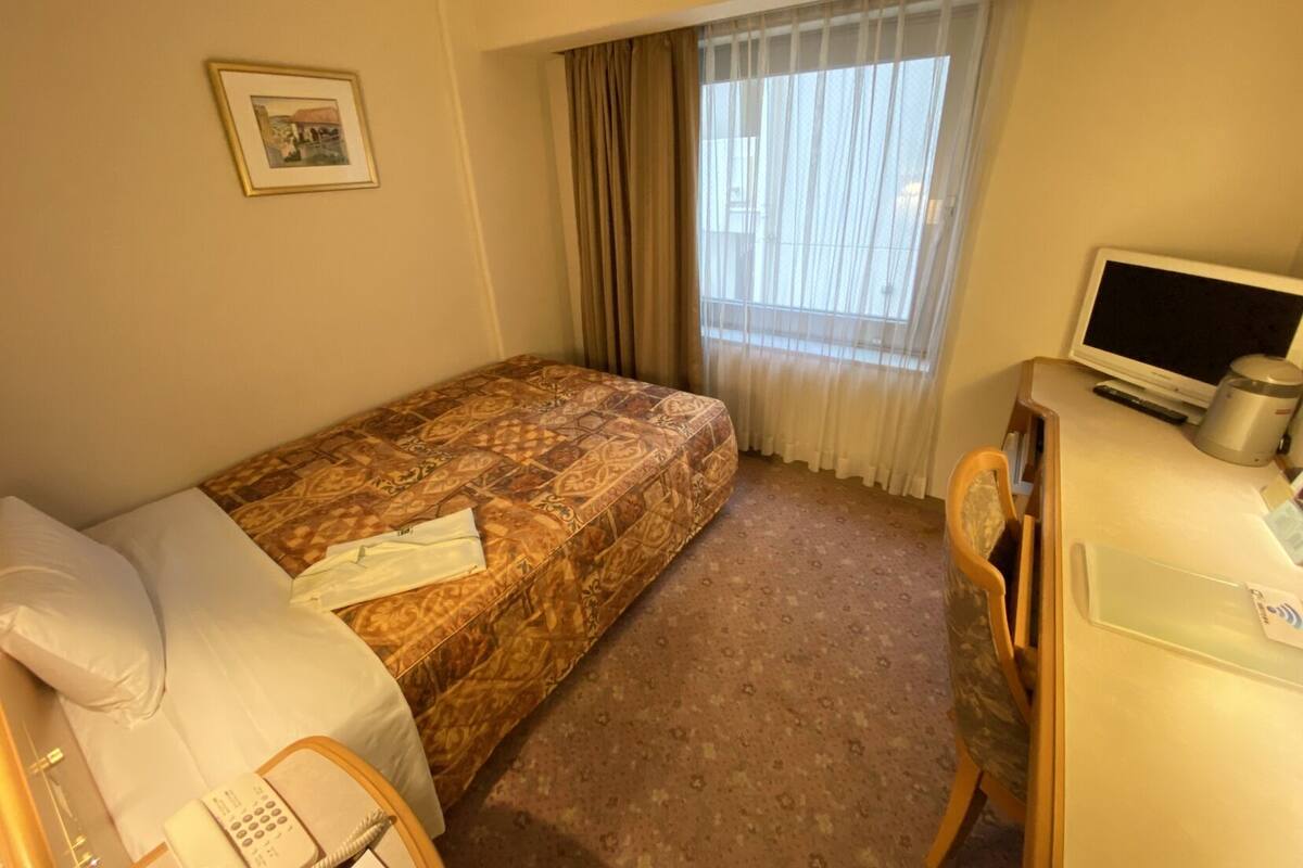 A hotel room with a bed, a desk with a computer, a chair, a window with curtains, and a framed picture on the wall.