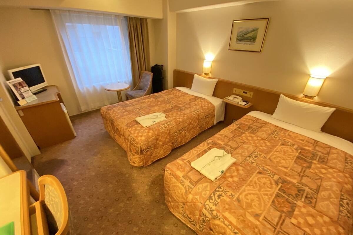A hotel room with two beds, a desk with a computer, a chair, a small table, and a window with curtains.