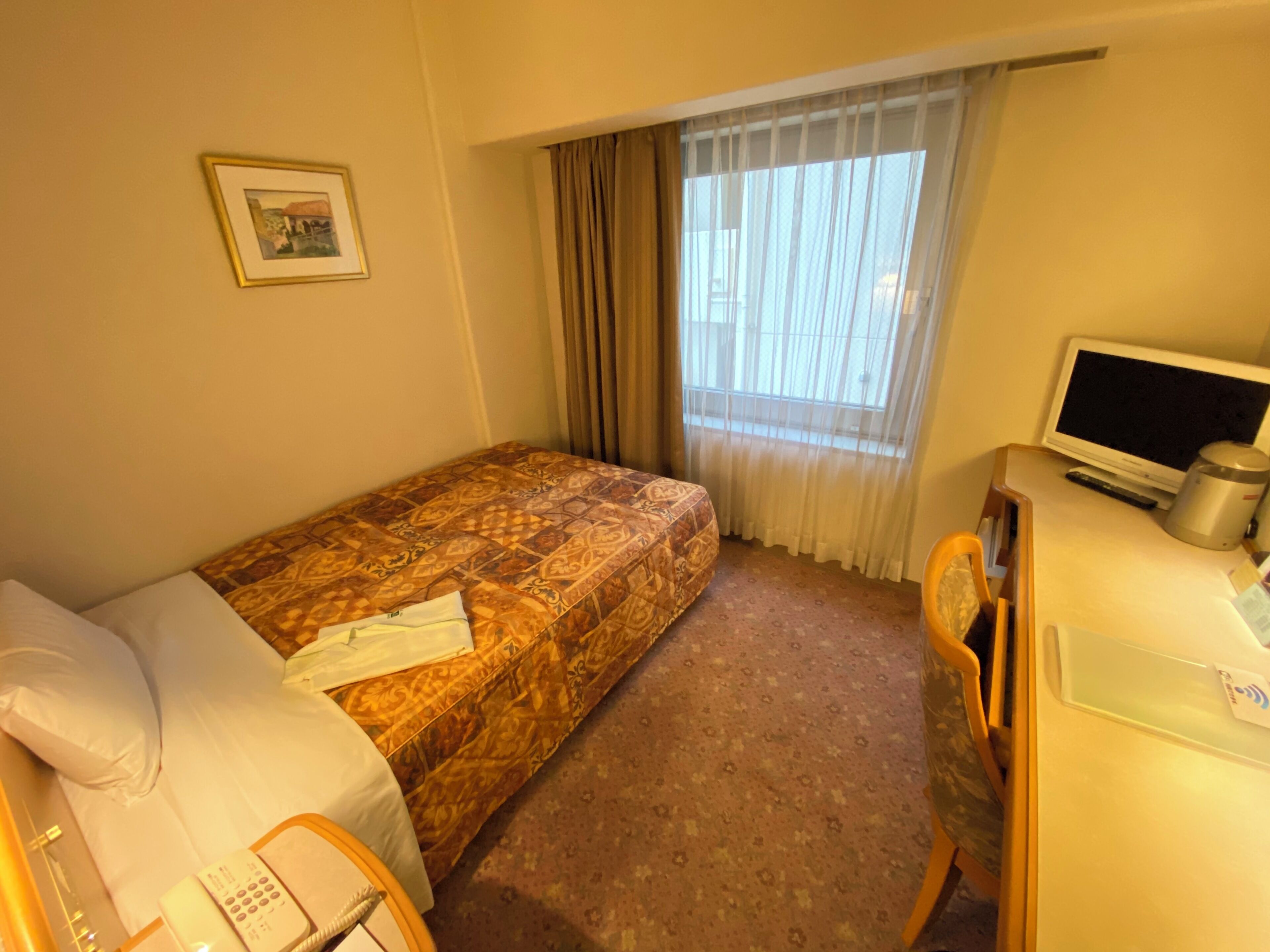 Habitación de hotel con cama, escritorio con ordenador, silla, teléfono y ventana con cortinas.