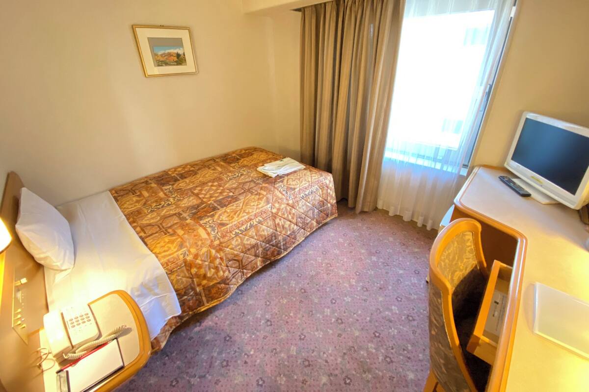 A hotel room with a bed, a desk with a television, a telephone, and a window with curtains.