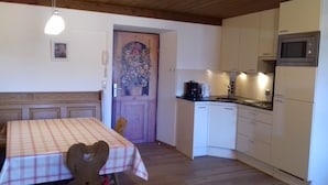 A small kitchen with white cabinets, a microwave, and a coffee maker. There is a table with a plaid tablecloth and a chair. A wooden door with a floral design is visible in the background. - Cosy Apartment In The Center Of Krimml, A Stroll Away From Krimml Waterfalls (Krimml)