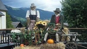 A rural outdoor scene with two statues of men, hay bales, sunflowers, and a wooden cart. - Cosy Apartment In The Center Of Krimml, A Stroll Away From Krimml Waterfalls (Krimml)