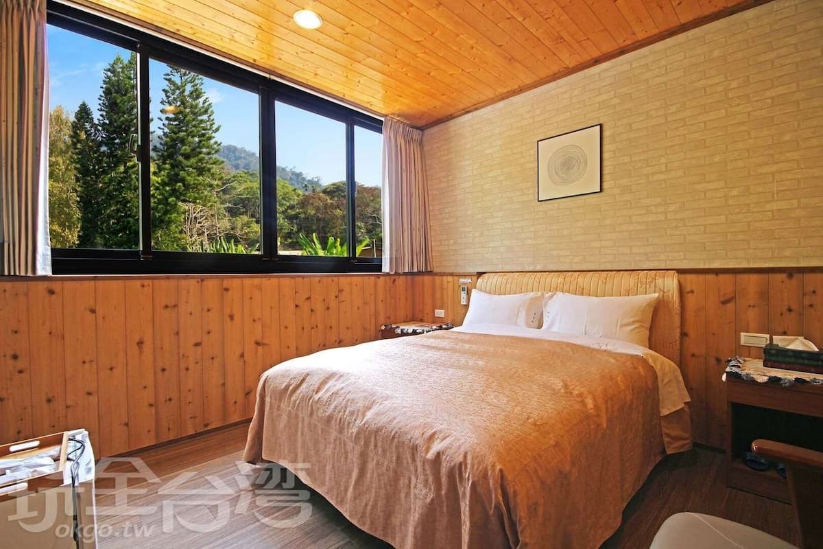 A bedroom with a large bed, wooden walls, a window with a view of trees, and a small bedside table.