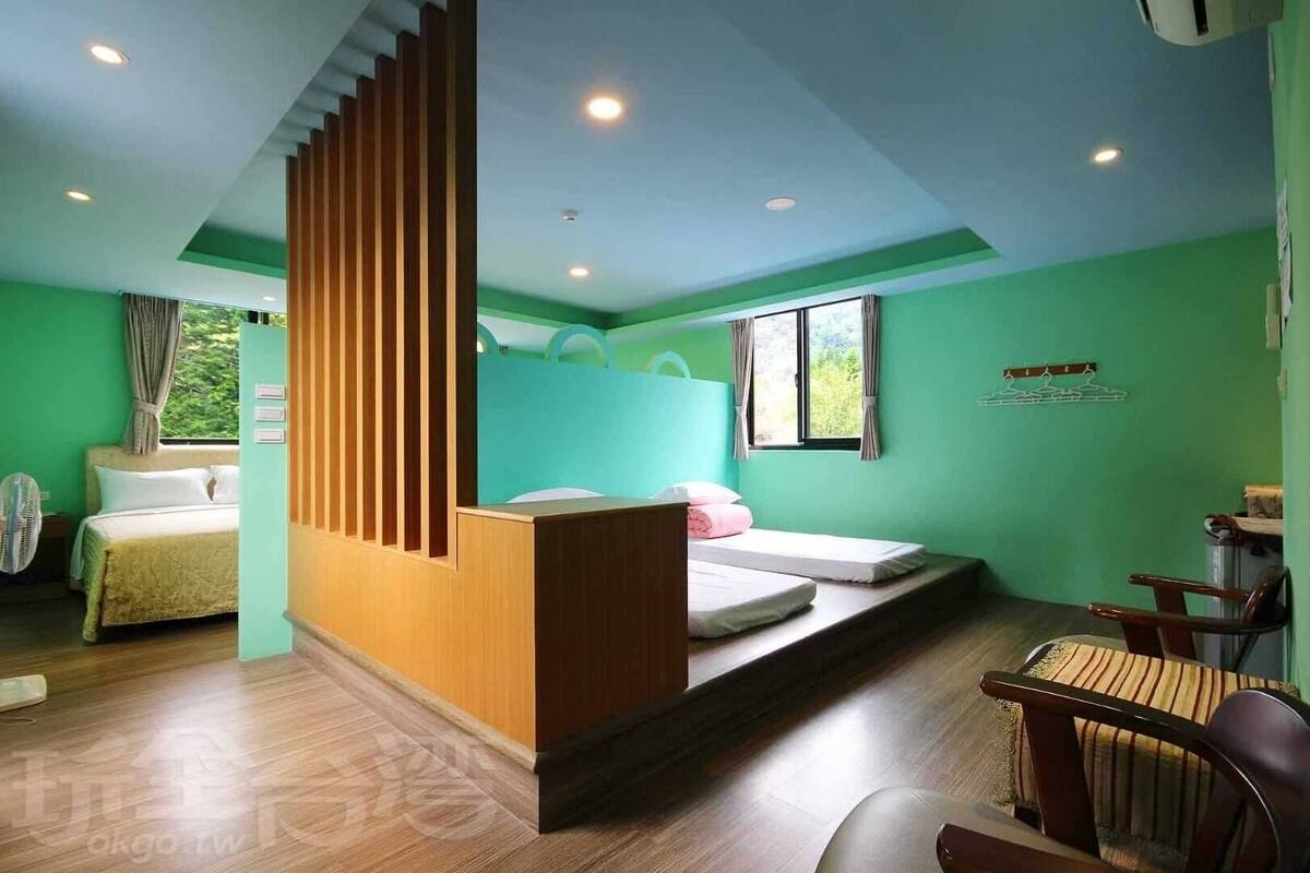 A room with a bed, a desk, and a chair. There is a window with curtains, a fan, and a wall-mounted shelf.