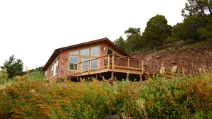 A house with a wooden deck and large windows, surrounded by dense vegetation. - Red Rock Overlook: A secluded mountain home high above the Uncompahgre River (Ridgway)