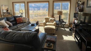 A living room with a sofa, armchair, and coffee table, large windows with a mountain view, and a bookshelf. - Red Rock Overlook: A secluded mountain home high above the Uncompahgre River (Ridgway)