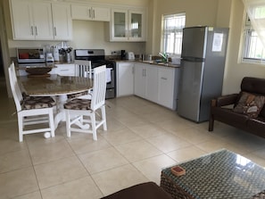 A modern kitchen with white cabinets, a stainless steel refrigerator, a dining table with chairs, and a brown leather sofa.