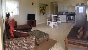 A living room with a sofa, a coffee table, and a television. There is a dining area with a table and chairs. The kitchen is visible with appliances and cabinets. - South Sea House Luxurious Apt No 1 (Cap Estate)