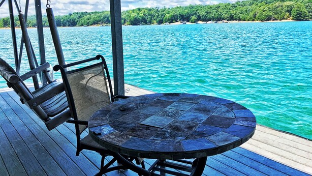 A wooden deck with a round table and chairs overlooking turquoise water and a forested shoreline. - Spacious, Family-Friendly Lake House Retreat w/ a Private Large 2-Story Dock (Cumming)