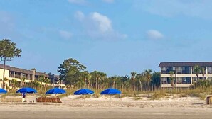 A beachside hotel with multiple blue umbrellas, a sandy beach, and a building with a flat roof and multiple windows. - Coastal King Couples Retreat Oceanfront Beach Property Convenient to Everything! (Hilton Head Island)