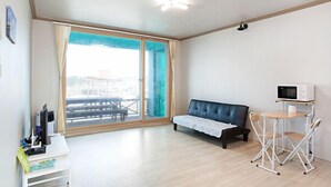 A modern room with a sofa, a small table with a chair, a microwave, and a view through large sliding doors. - Jeju Sky Sea Pension (Jeju City)