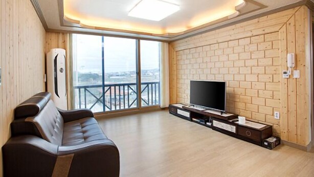 A modern living room with a large window, a flat-screen TV on a wooden stand, and a leather sofa. - Jeju Sky Sea Pension (Jeju City)