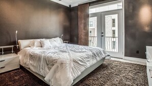 Ein Schlafzimmer mit einem Bett, einem Deckenventilator, einem Nachttisch, einer Kommode und einem großen Fenster mit Blick auf Gebäude.