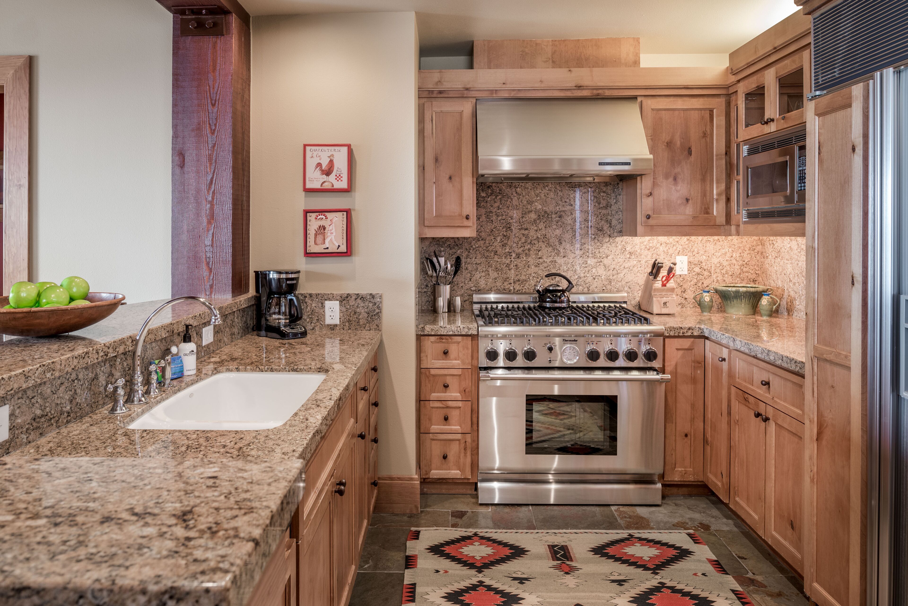 A kitchen with granite countertops, stainless steel appliances, wooden cabinets, and a patterned rug.