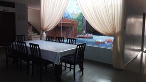 A dining area with a table and chairs, a view of a pool with a waterfall, and curtains on the windows. - ArRayyan Guesthouse & Homestay (Taiping)