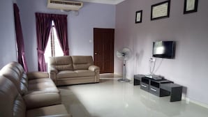 A living room with a beige sofa, a standing fan, a TV on a black stand, and a door in the background. - ArRayyan Guesthouse & Homestay (Taiping)
