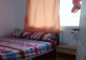 A single bed with a colorful quilt, a bedside table with a remote control, and a window with a curtain. - Natcha Place Rangsit Donmuang (Lam Luk Ka)