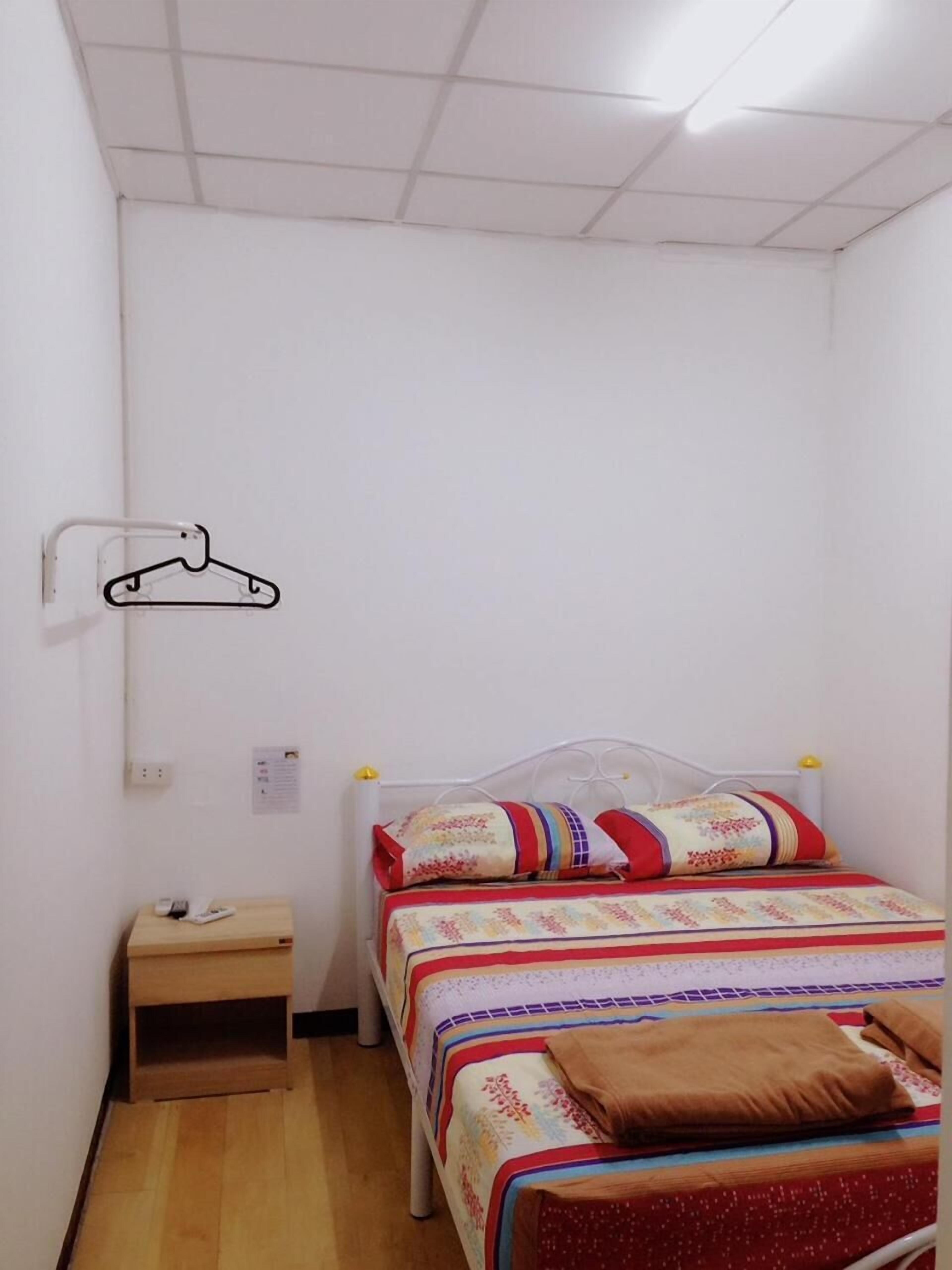 Quarto pequeno, com uma cama de solteiro, mesa de cabeceira de madeira, cabideiro e roupa de cama estampada.