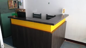 A reception desk with a dark wood finish and a yellow accent. There are two chairs behind the desk, and a small sign with a picture of a dessert is on the wall to the right. - Natcha Place Rangsit Donmuang (Lam Luk Ka)