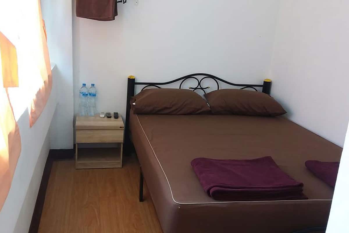 A single bed with a headboard, a wooden nightstand with a lamp, two bottles of water, and a small table with a calculator.