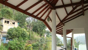 Un espace extérieur abrité, doté d’une structure de toit en bois, de colonnes apparentes et offrant une vue sur une colline parsemée de bâtiments et de végétation.