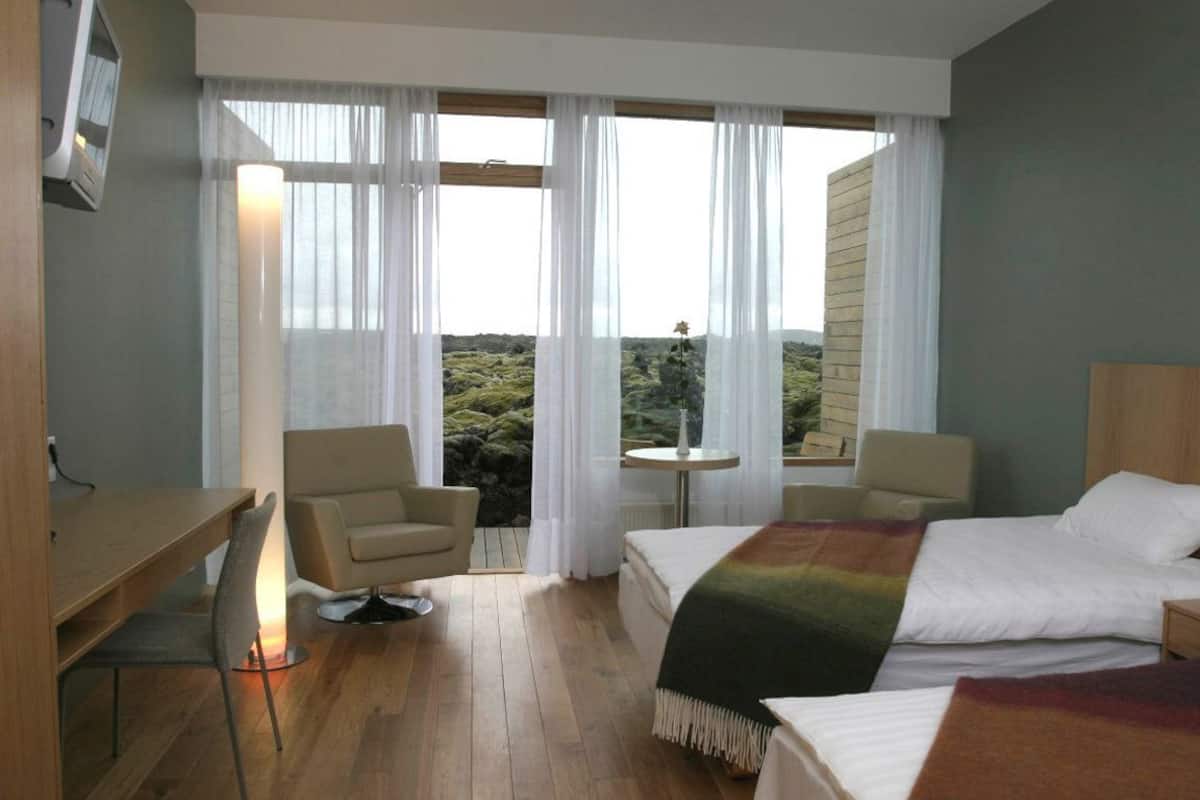 A hotel room with a bed, two chairs, a small table, and a view of a mountainous landscape through large windows.