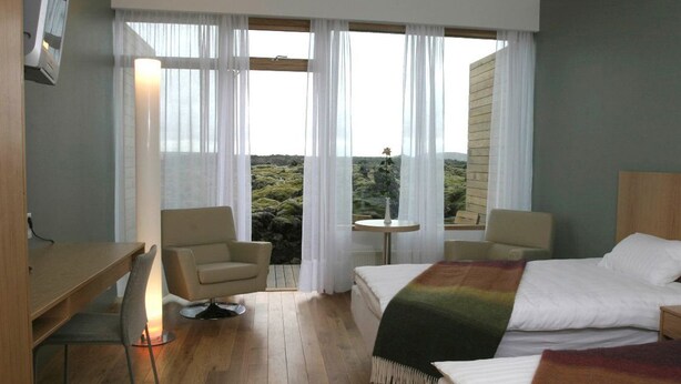 Une chambre d’hôtel avec un lit, deux chaises, une petite table et une vue sur un paysage montagneux grâce à de grandes fenêtres.