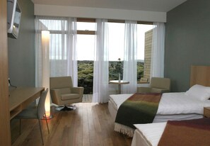 Een hotelkamer met een bed, twee stoelen, een kleine tafel en uitzicht op een bergachtig landschap door grote ramen.