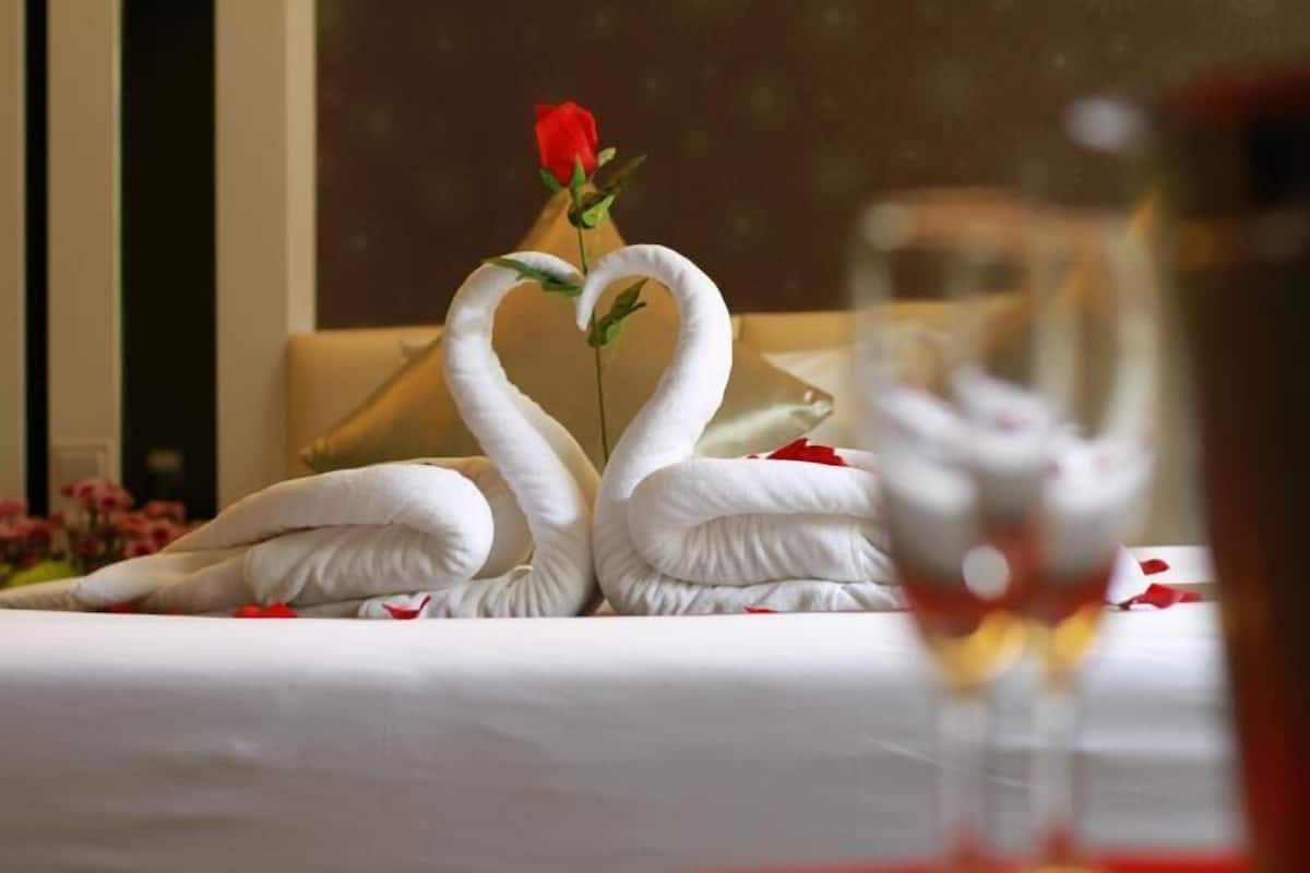 A bed with a rose and two swan-shaped pillows, a glass of wine, and a vase with a single red rose.