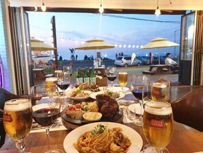 A dining table with multiple glasses of beer, plates of food, and a view of the outdoors.