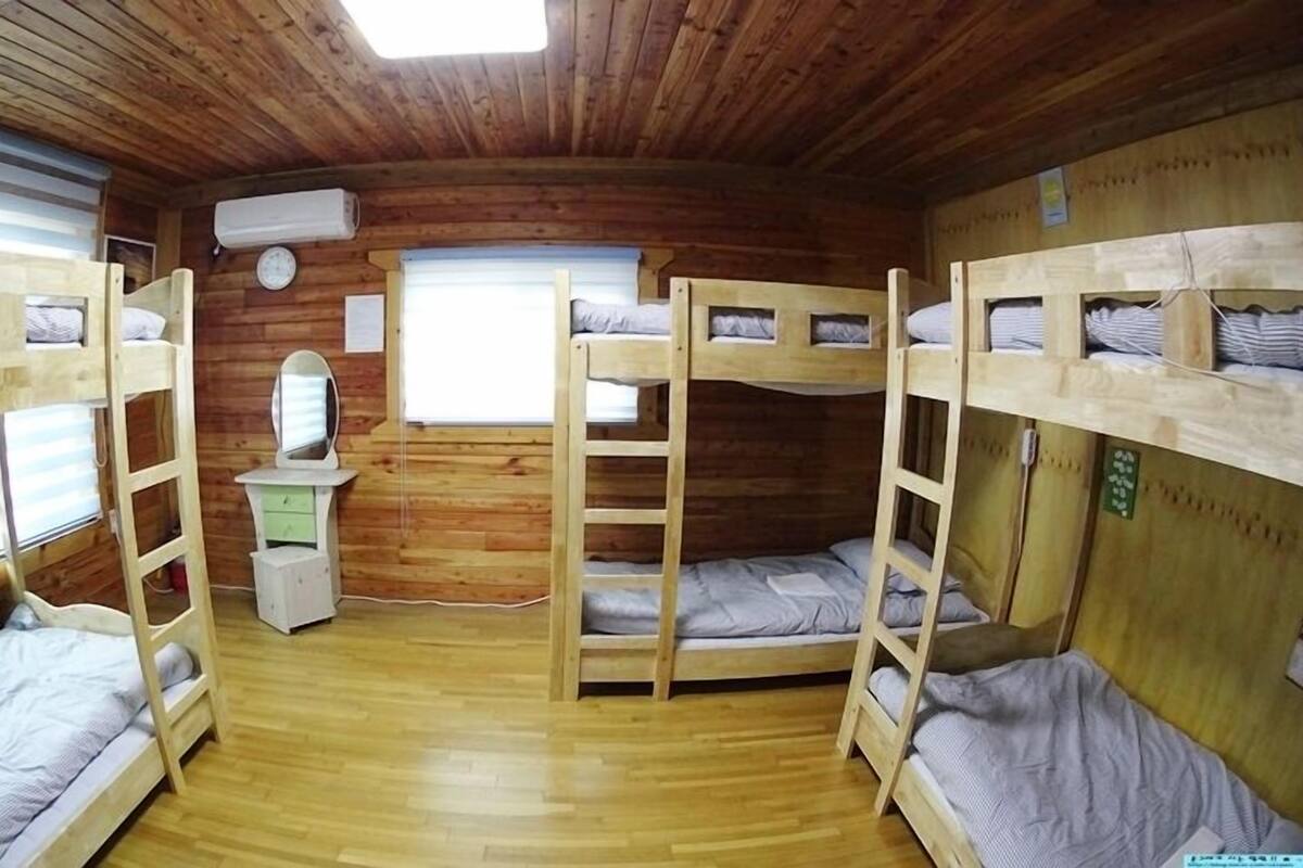 Eine Holzhütte mit Etagenbetten, einem kleinen Schminktisch mit Spiegel, einer Uhr und einem Fenster mit Jalousien.