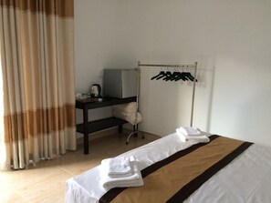 A hotel room with a bed, a desk with a kettle, a clothes rack, and a window with curtains.