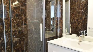 Une salle de bain moderne avec une douche à parois de verre, un meuble-lavabo blanc doté d’un grand miroir et des murs recouverts de marbre foncé.