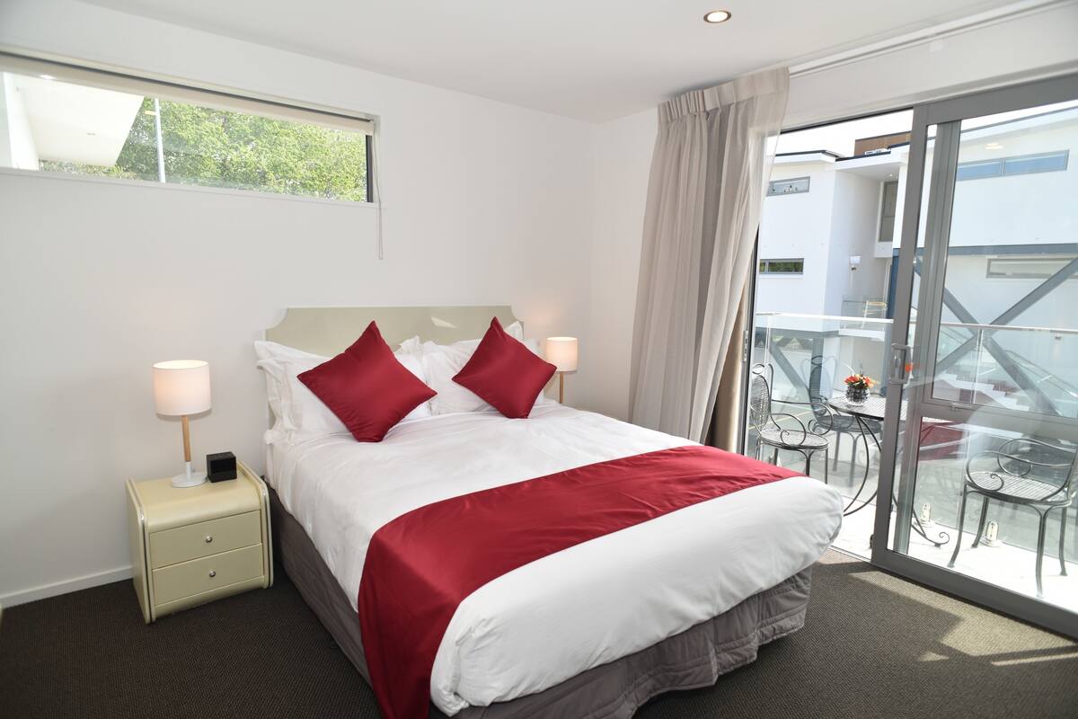 A hotel room with a bed, two bedside tables, a window, and a balcony with outdoor furniture.