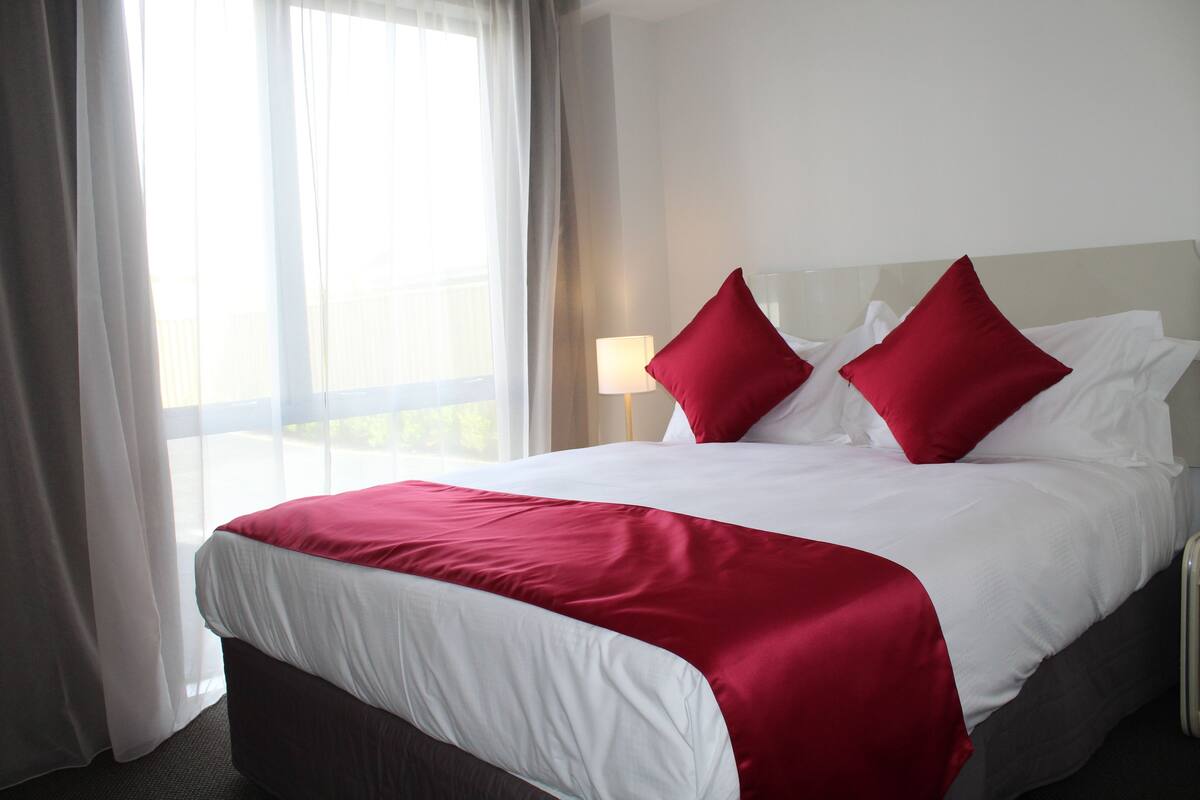 A hotel room with a large bed, white and red bedding, a bedside lamp, and a view of the outdoors through sheer curtains.