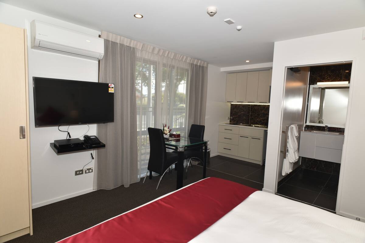 A hotel room with a bed, a desk with a chair, a TV, a kitchenette, and a bathroom.