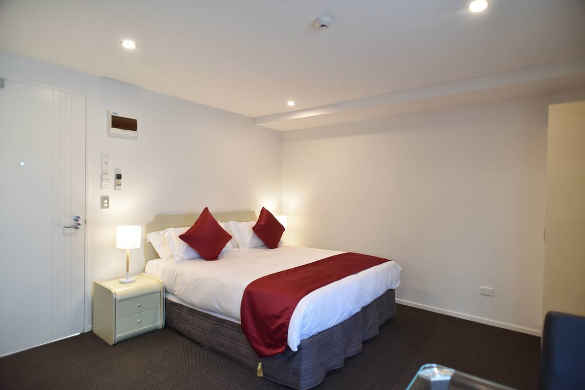 A hotel room with a bed, a nightstand, a lamp, a door, and a wall-mounted thermostat.