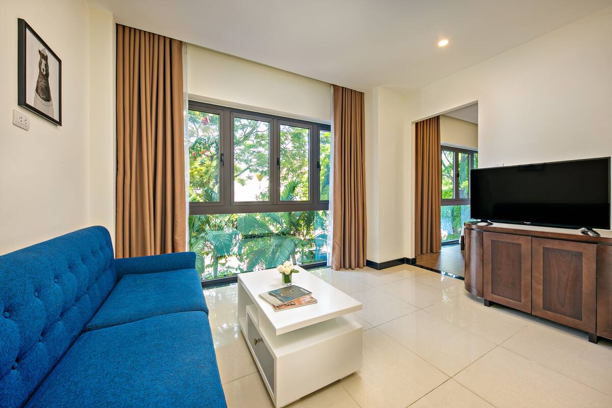 A modern living room with a blue sofa, a white coffee table with a book and vase, a TV on a wooden cabinet, and large windows with curtains.