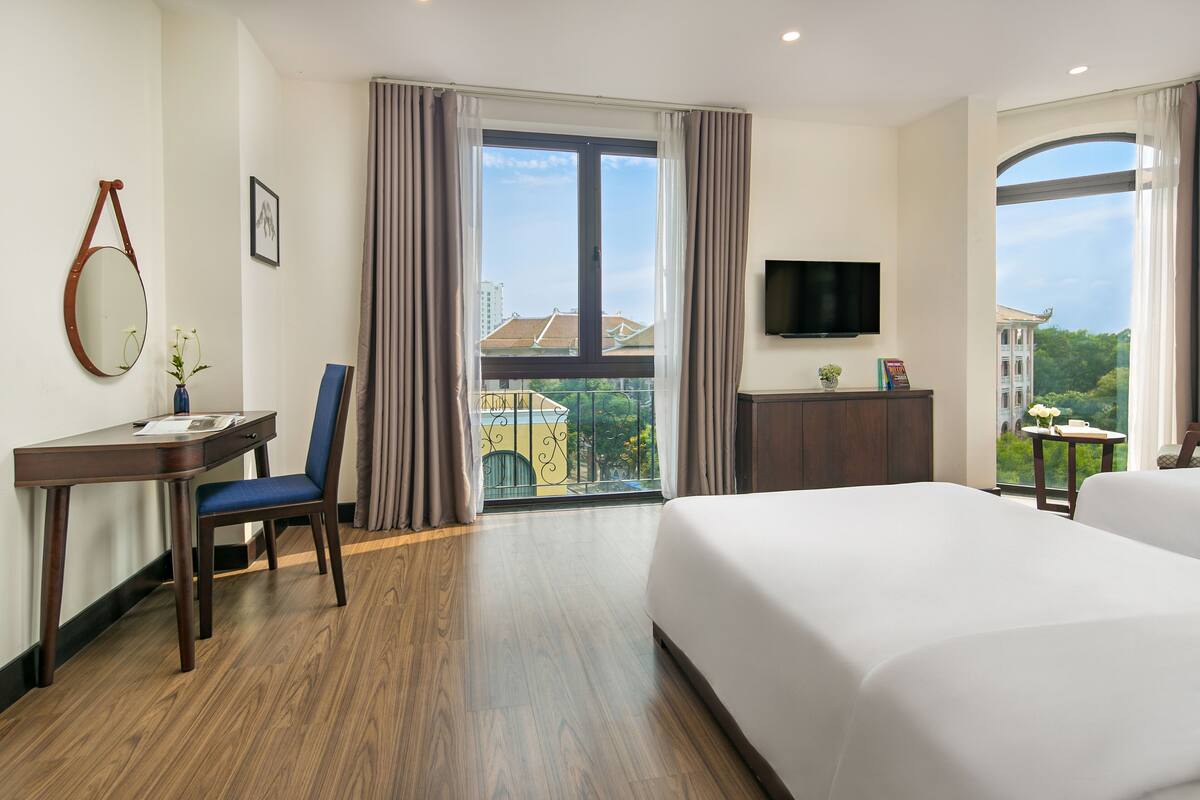 A hotel room with a large bed, a desk with a chair, a TV mounted on the wall, and a balcony with a view of buildings.