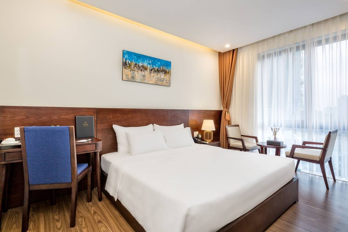 A hotel room with a large bed, a desk with a computer, a chair, a small table with a lamp, and a painting on the wall.