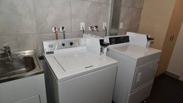 Une buanderie équipée de deux machines à laver et deux sèche-linge blancs, d’un évier en acier inoxydable et d’un mur recouvert de carreaux.