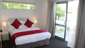 A hotel room with a large bed, two nightstands, a window with a view of greenery, and a sliding glass door to an outdoor area.