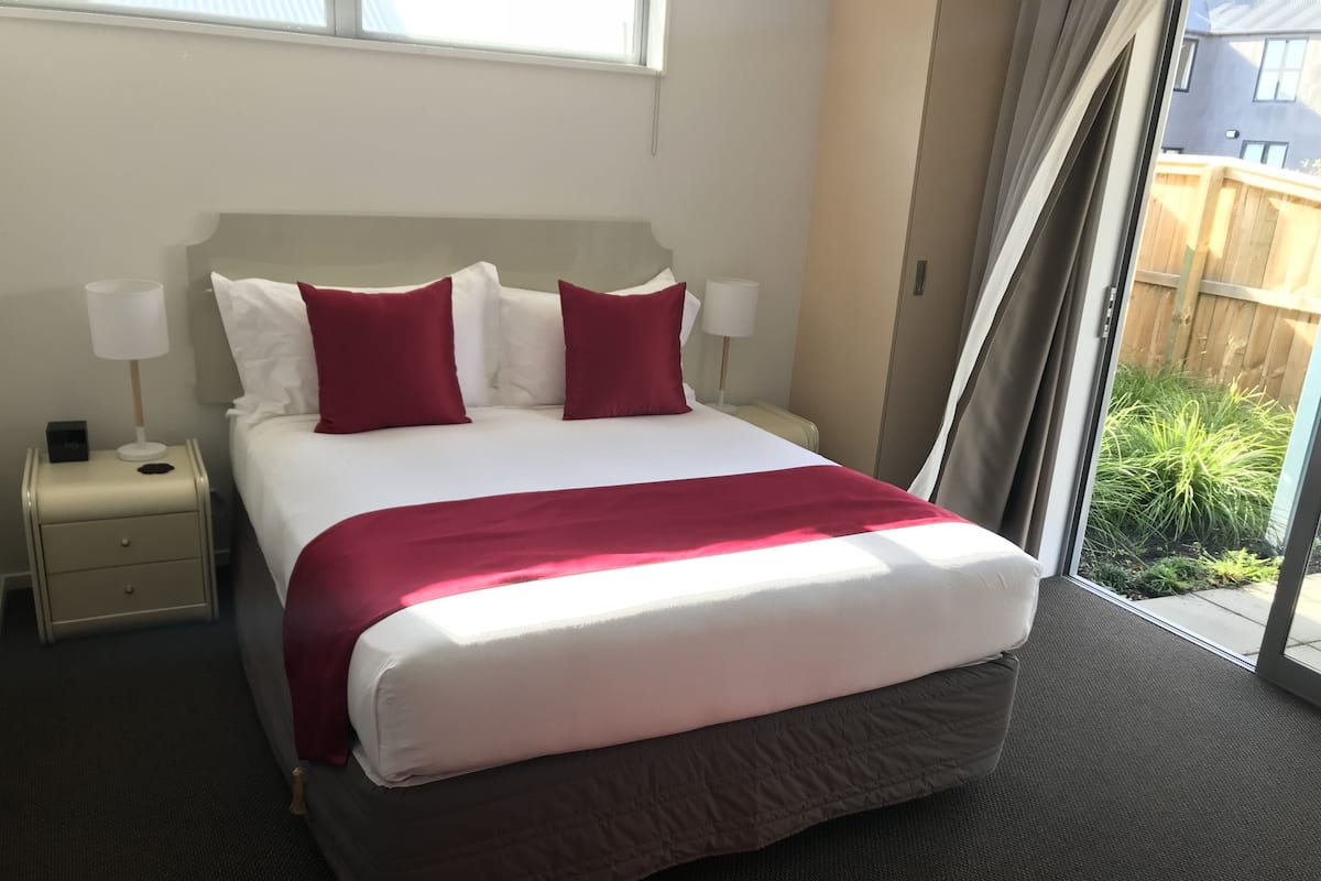 Hotellihuone, jossa on suuri sänky, kaksi punaista tyynyä, valkoinen pääty, yöpöytä lampun kanssa ja näkymä puuaidalle ja viheralueelle.