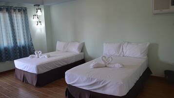 Deux lits simples avec du linge de lit blanc et des couvre-lits foncés, un plancher en bois, un rideau et un luminaire fixé au mur.