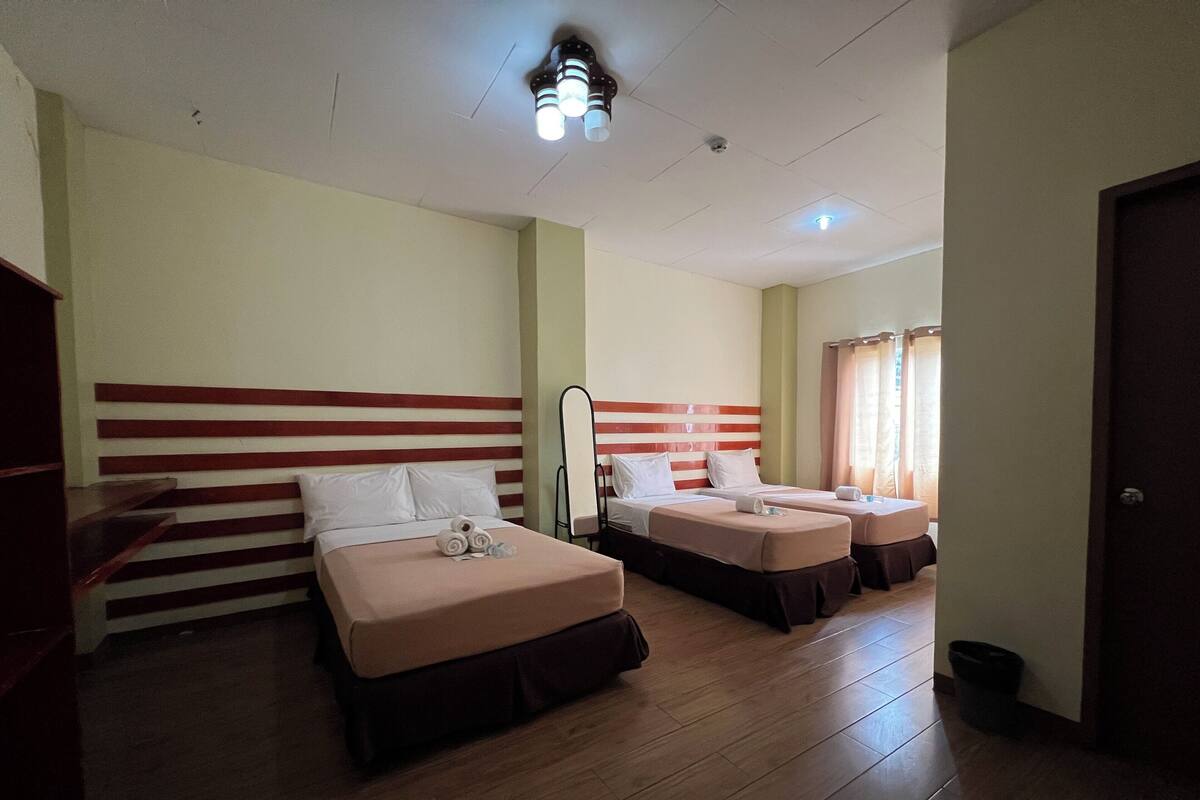 Habitación de hotel con tres camas, piso de madera y techo con detector de humo e iluminación.