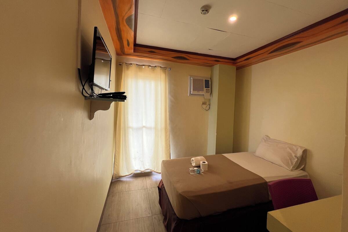 Una habitación de hotel con cama, televisión montada en la pared, ventilador de techo y ventana con cortinas.