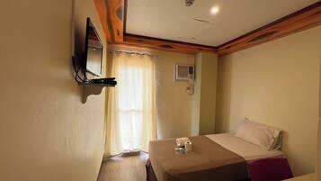 Ein Hotelzimmer mit einem Bett, einem an der Wand befestigten Fernseher, einem Deckenventilator und einem Fenster mit Vorhängen.