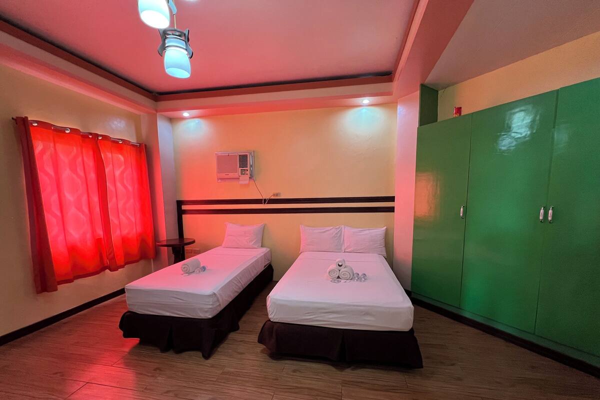Una habitación de hotel con dos camas individuales, un armario verde, una lámpara en el techo y un aire acondicionado instalado en la pared.