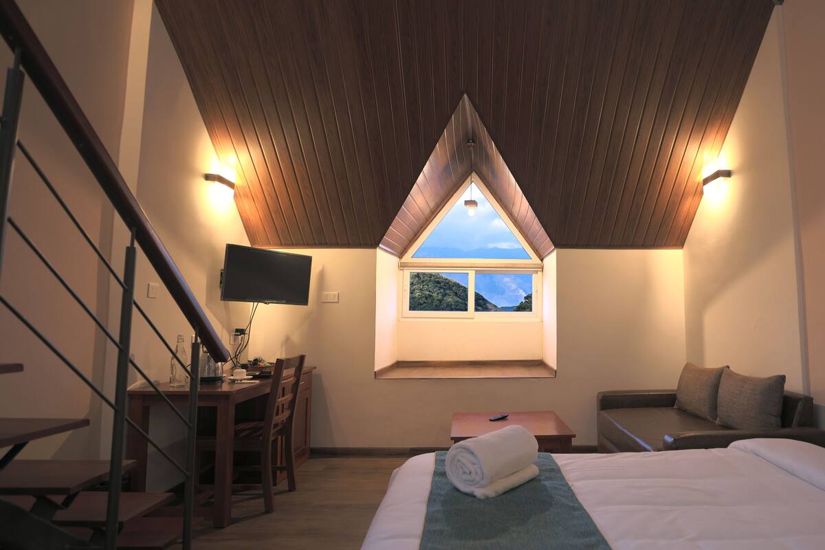 Ein Schlafzimmer mit schräg verlaufender Decke, einem Bett, einem Schreibtisch mit Stuhl, einem kleinen Tisch und einem Fenster mit Blick auf eine Hügel.